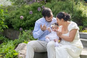 A couple in white attire sits outdoors in a garden, smiling and playing with their baby girl on a sunny day.