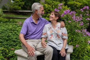 Elderly couple sitting on a stone bench in a garden, smiling at each other surrounded by purple flowers.