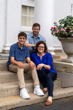 Family of three smiling on outdoor steps near white columns and pink flowers on a sunny day.