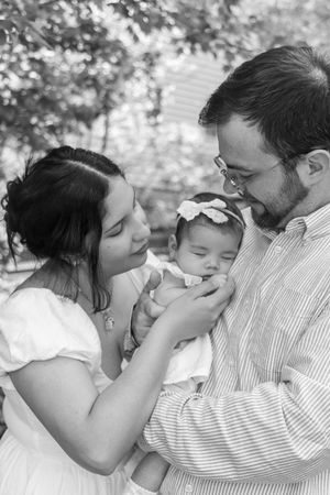 Black and white photo of parents lovingly gazing at their newborn baby girl with a bow headband outdoors.