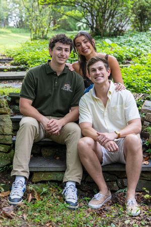 Three smiling young adults posing outdoors on stone steps surrounded by lush green garden foliage.