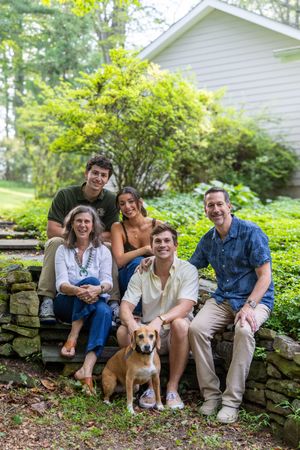 Family of five poses with a beagle dog on stone steps surrounded by lush green trees near a white house.