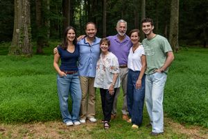 Six smiling family members pose together outdoors in a lush green forest setting for a casual group portrait.