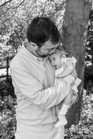 Black and white photo of a father tenderly holding his infant baby outdoors near a tree.