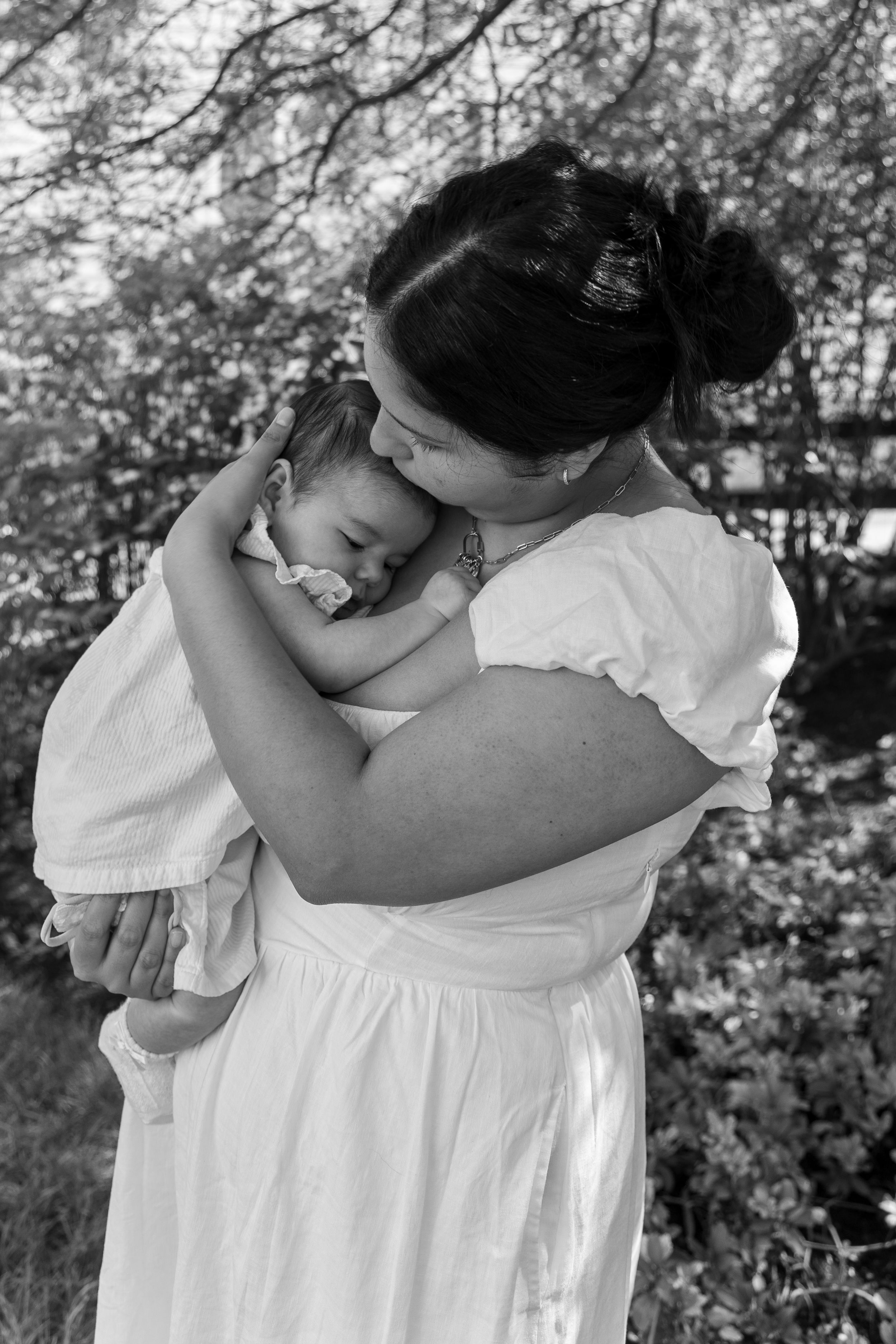 Black and white photo of a mother in white dress tenderly holding her sleeping newborn baby outdoors.