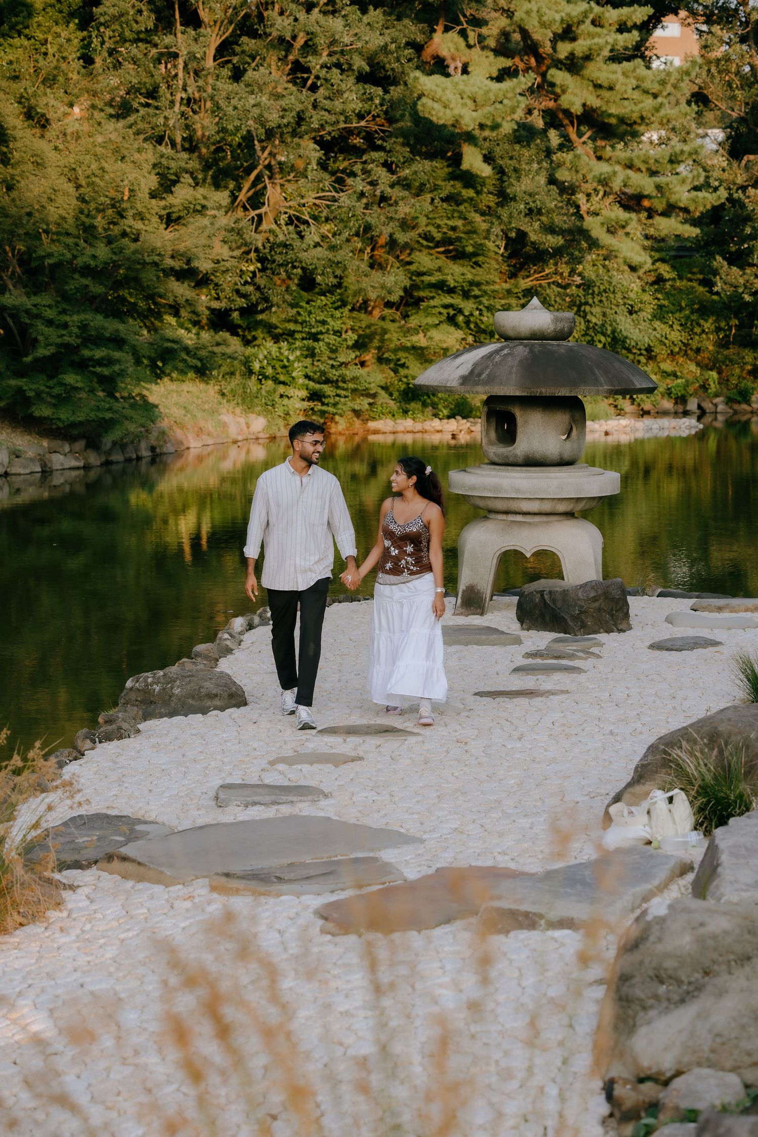 A couple walks hand in hand along a serene Japanese garden path beside a tranquil pond with traditional stone lantern.