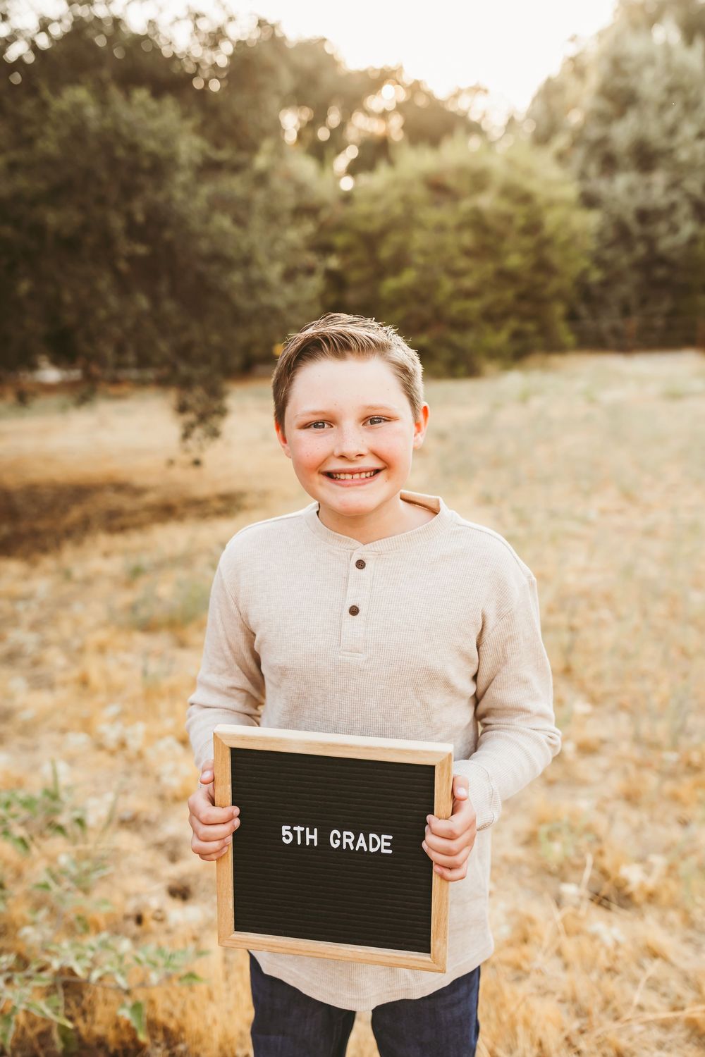 A student holds up a black letter board displaying '5th Grade' in an outdoor setting.