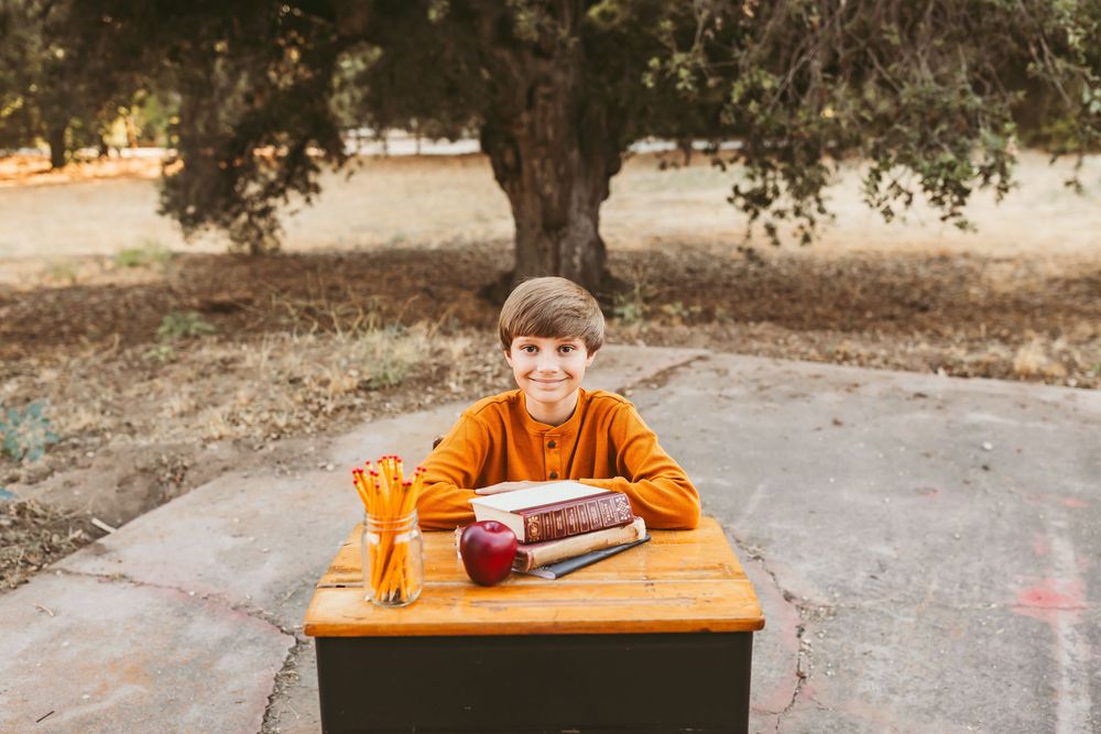 A person in an orange shirt sits at a wooden desk outdoors under trees on a sunny autumn day.