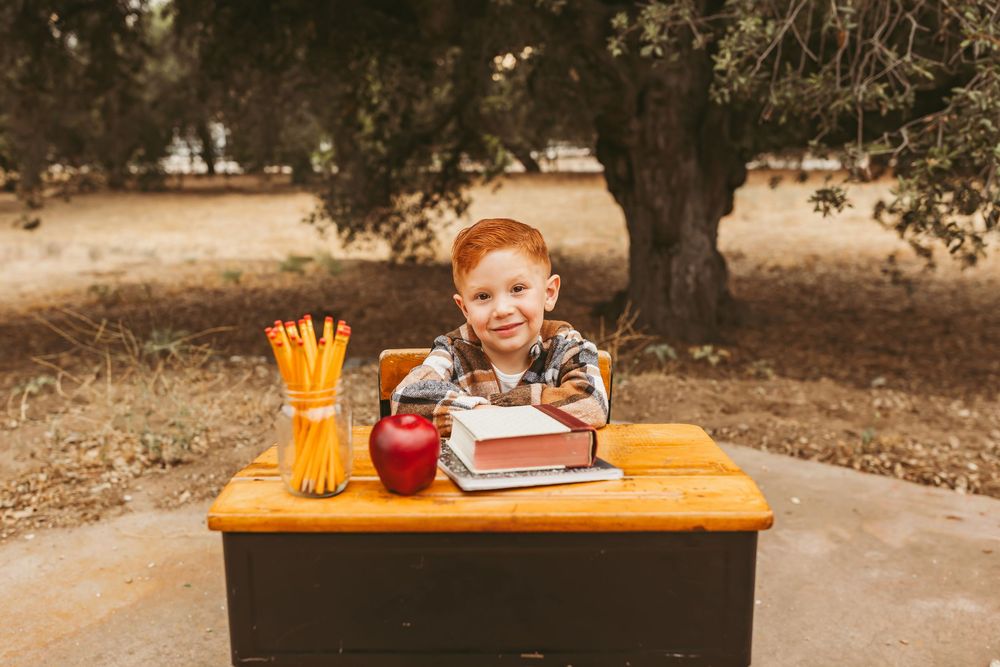 A young student sits at an orange table with school supplies and a red drink outdoors.