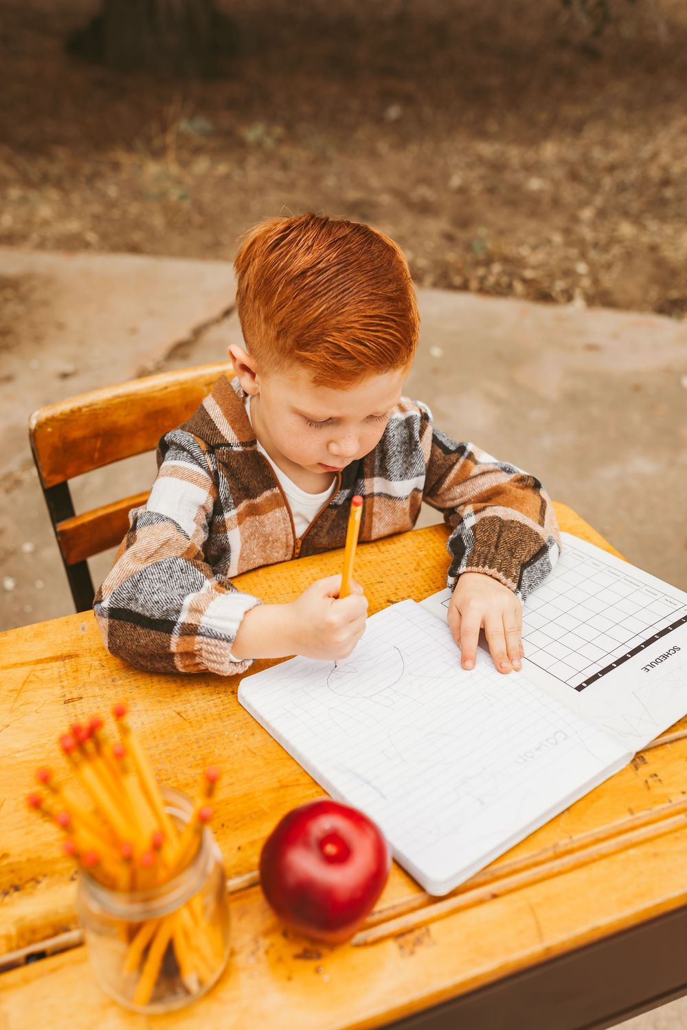 Student writes in notebook at wooden desk with red apple and orange flowers in foreground.
