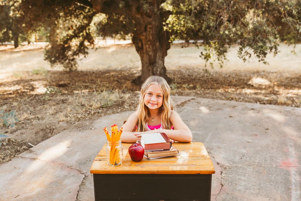 Student sitting at a wooden desk outdoors with school supplies and a letter board on a sunny day.