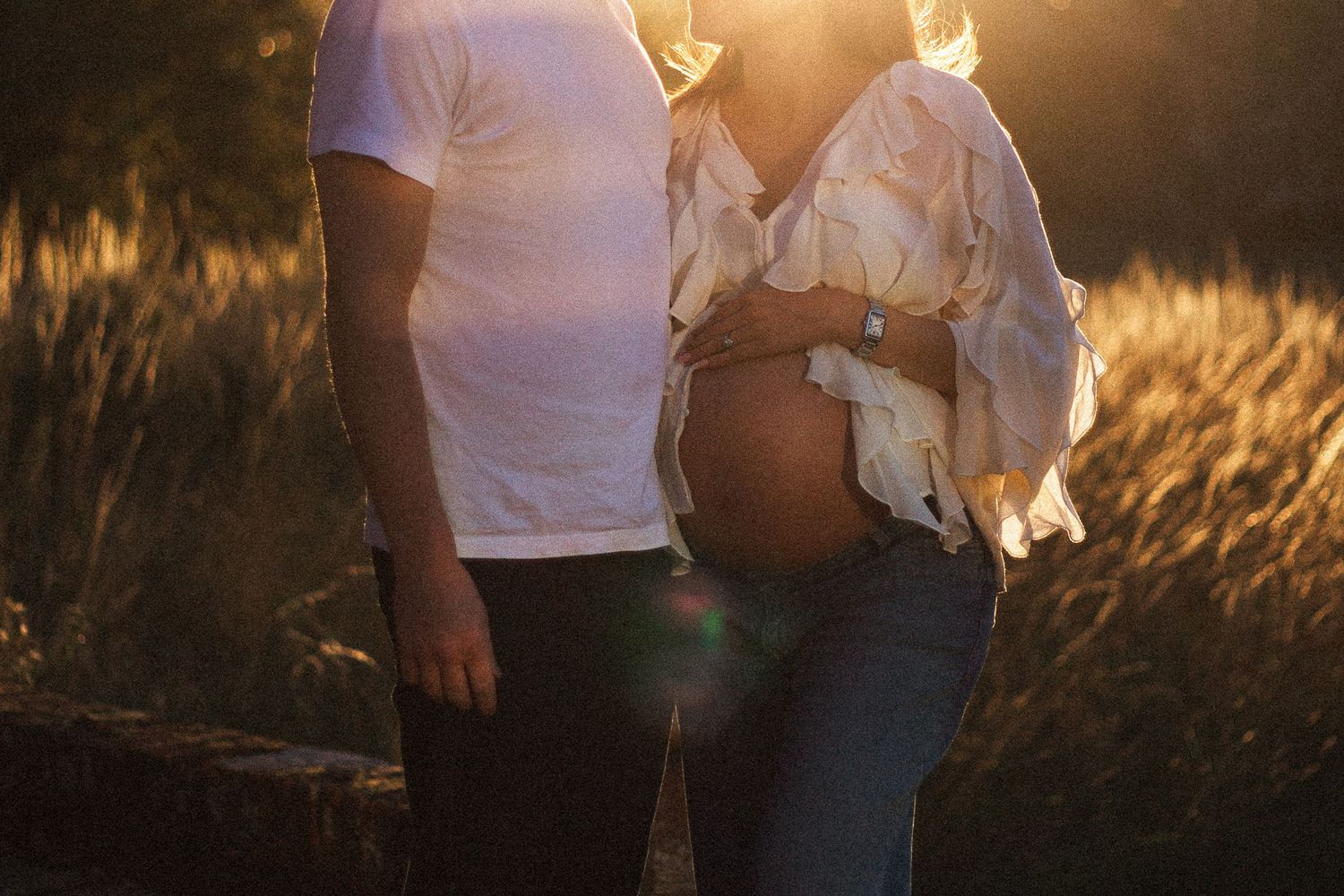 Pregnant woman standing in golden-hour light in Surrey, gently holding her bump whilst hugging partner — a natural, emotive maternity