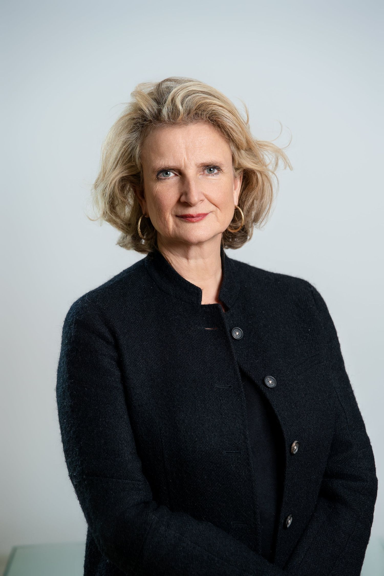 Professional portrait of a blonde woman in a dark jacket against a light gray background.