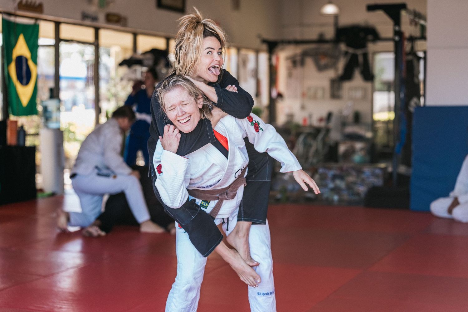 Students practice martial arts techniques in a training studio with mats and Brazilian flags on the wall.