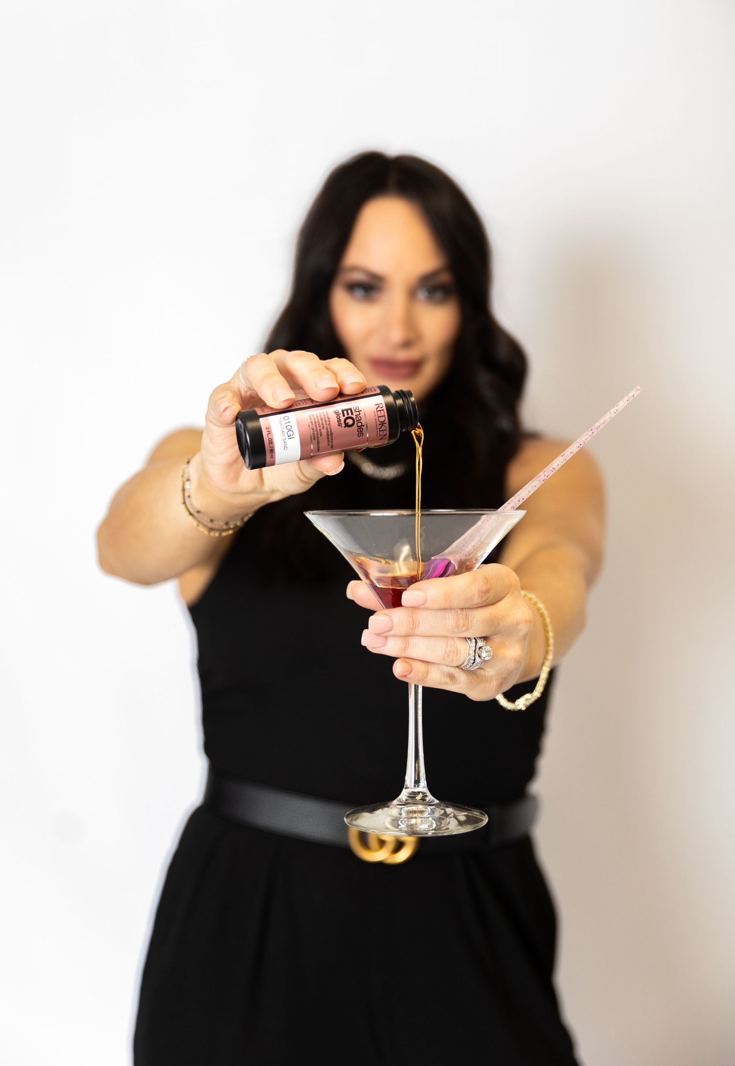 A person in a black dress pours a pink cocktail into a martini glass against a white background.