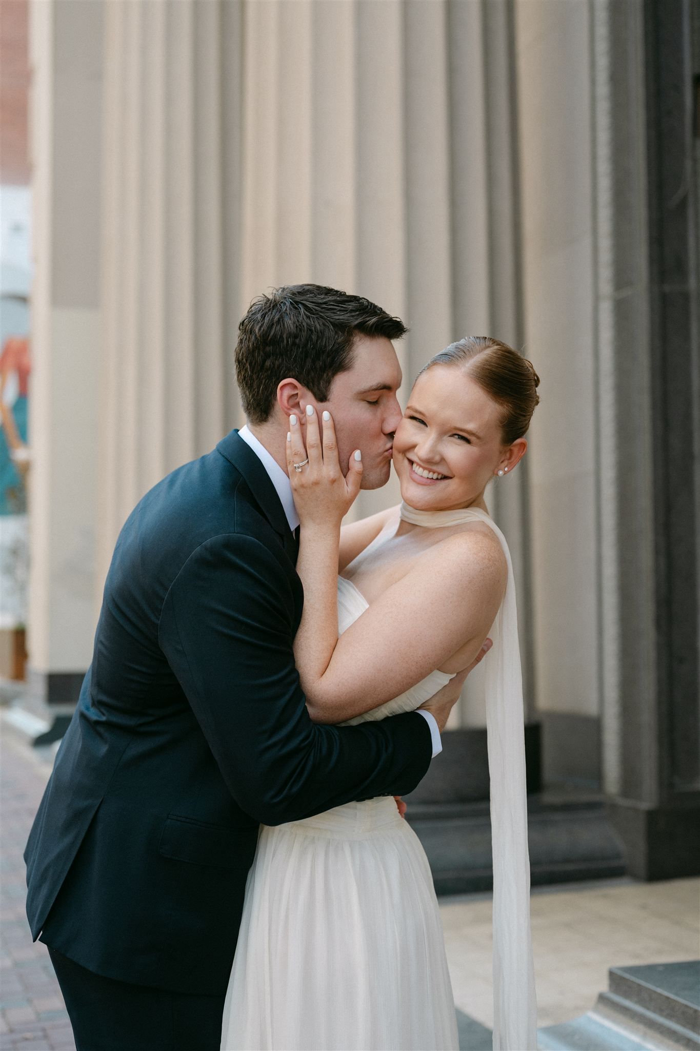 A bride and groom share a romantic kiss in front of white columns at their wedding venue.