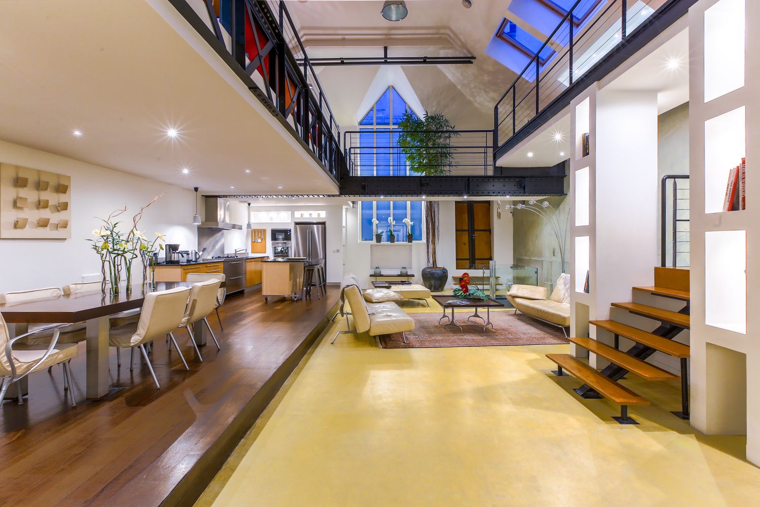 Spacious industrial loft with double-height ceilings, mezzanine level, open kitchen, and yellow floors.