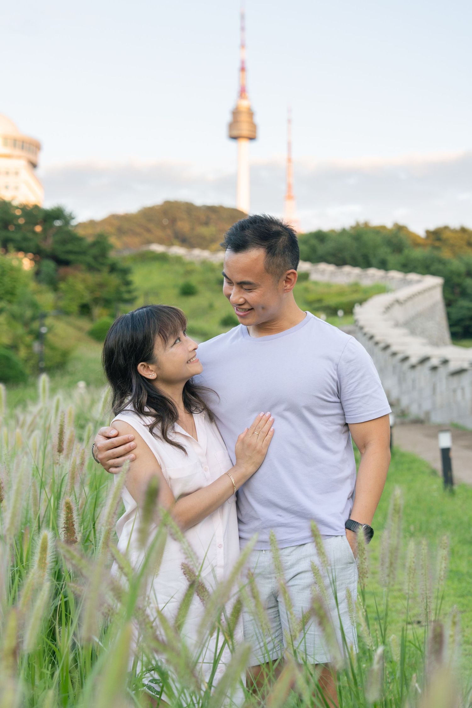A couple embraces in a grassy field with Seoul Tower visible in the background on Namsan Mountain.