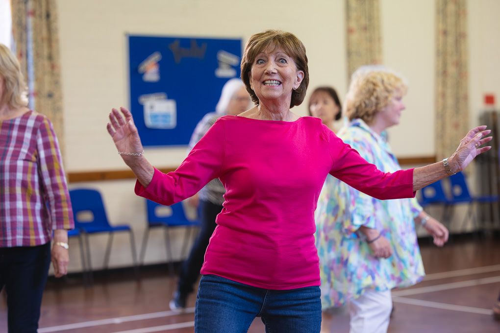 Seniors participating in a group dance class wearing pink and plaid shirts in a Hitchin community centre.