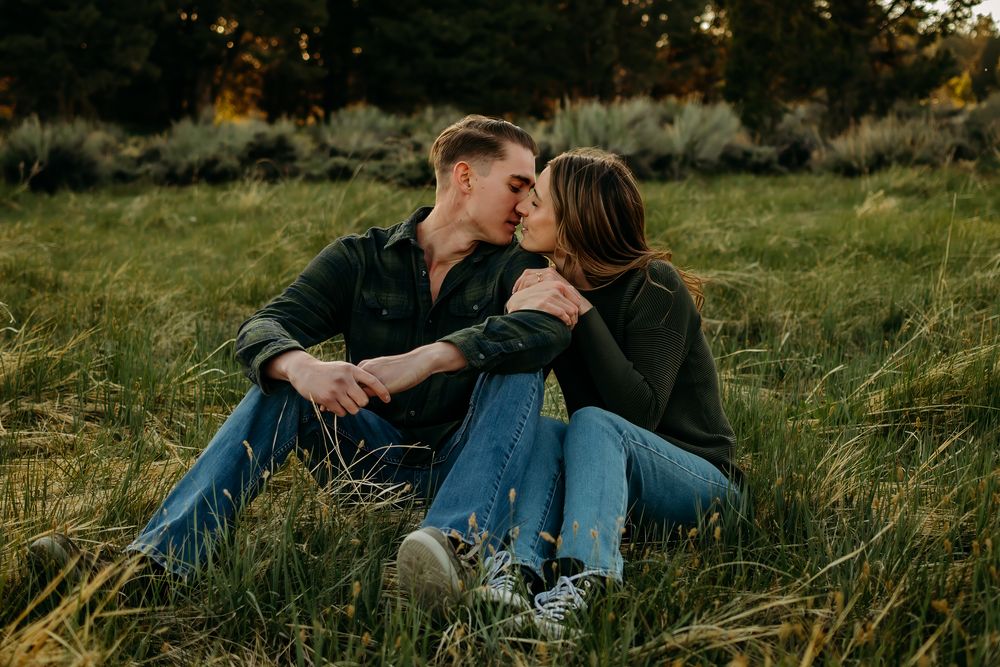 Couple sitting down and kissing in a meadow with trees behind them