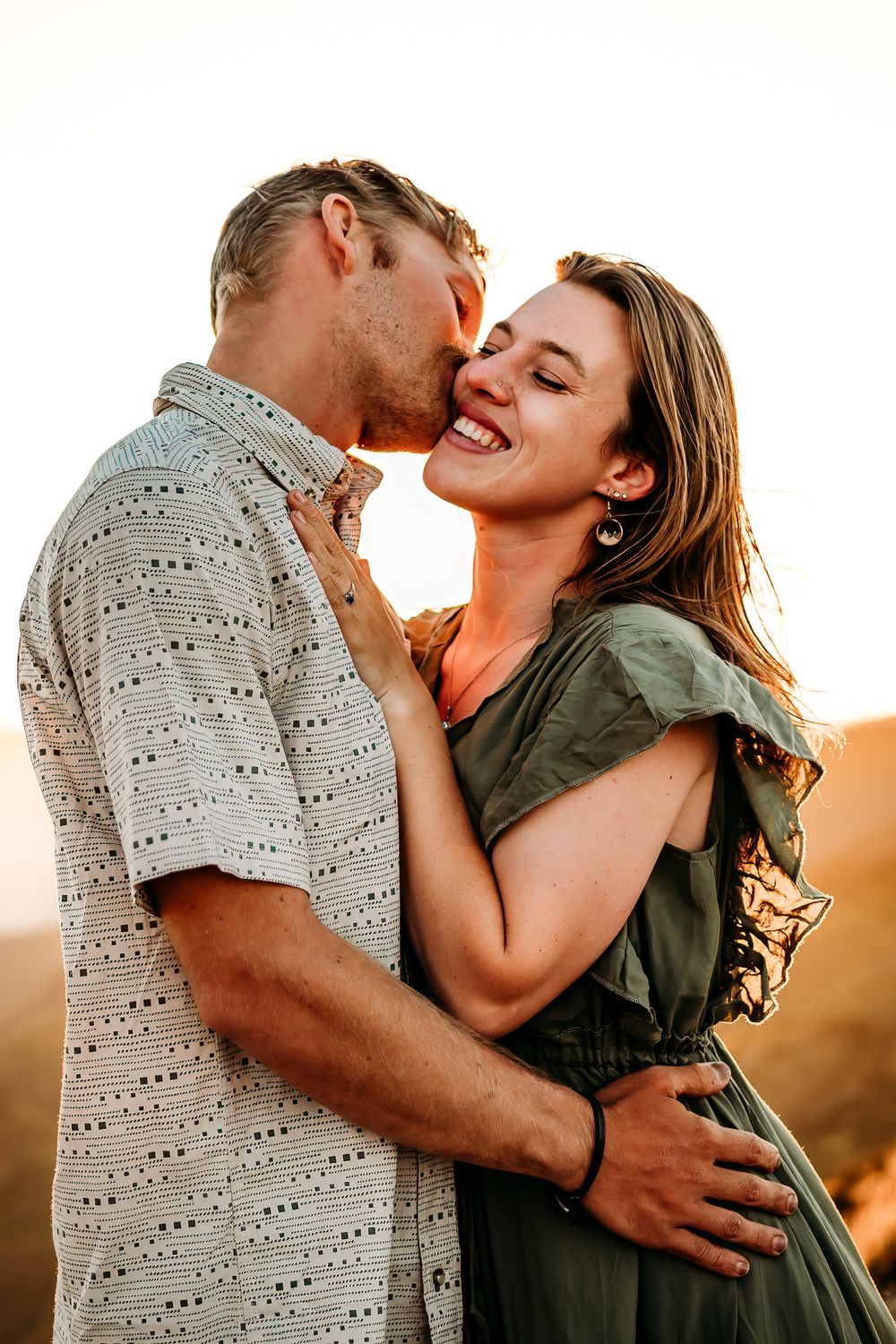 male kissing female on the cheek with mountains and the sun behind them.