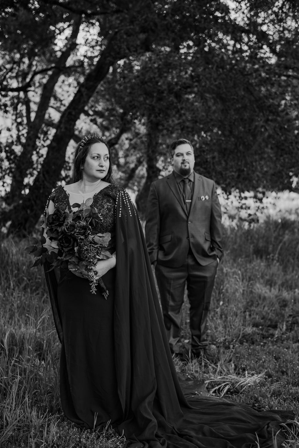Gothic-styled couple in black attire poses dramatically in a moody outdoor setting.