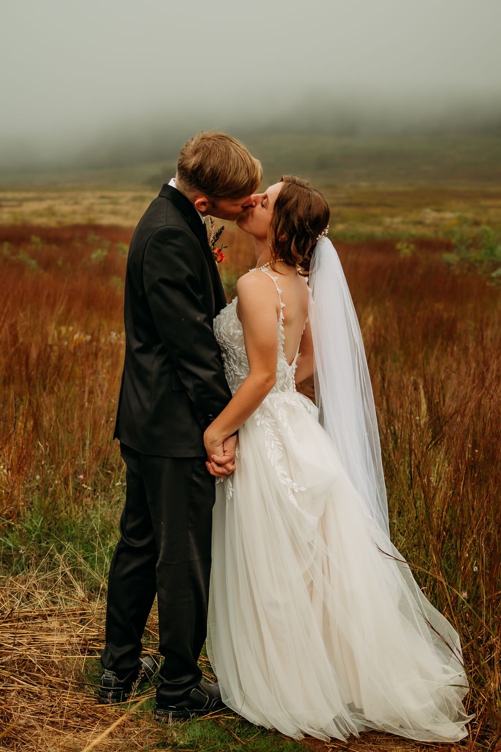 Intimate wedding moment captured in a misty field with flowing white dress and veil.