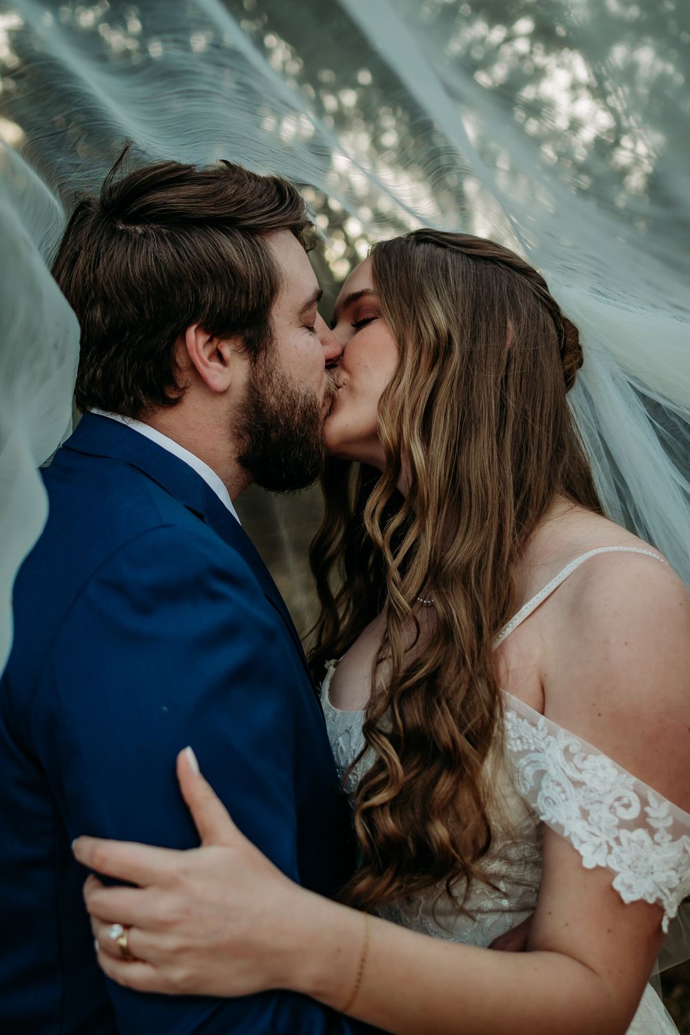 An intimate wedding kiss captured under flowing veil and dappled sunlight.