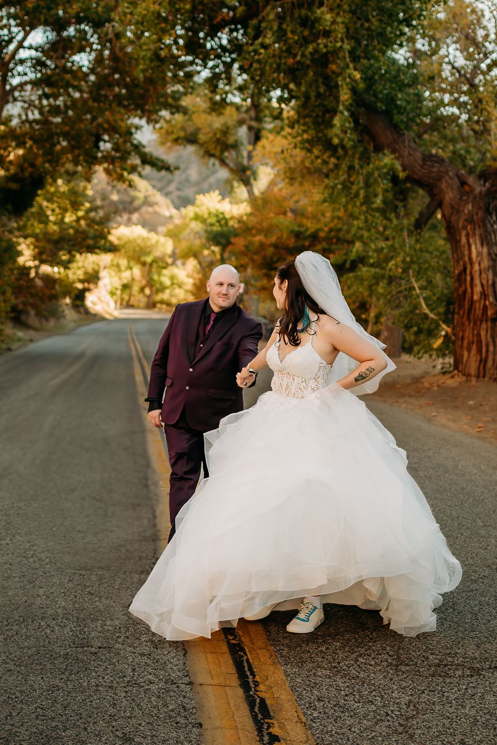 Couple in wedding attire poses playfully on yellow lined road surrounded by autumn trees.