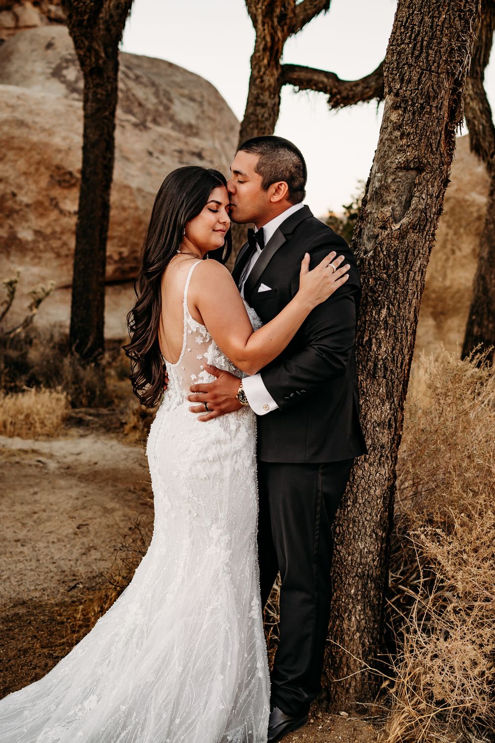 A romantic wedding portrait of a couple kissing beneath rustic trees.