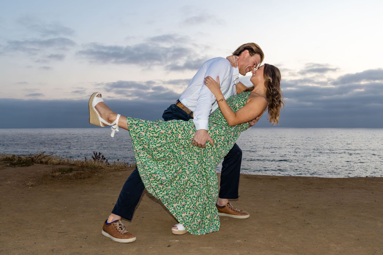 San Diego couple portrait photography of a man playfully dipping a woman in a green dress on a cliff overlooking the ocean at sunset.