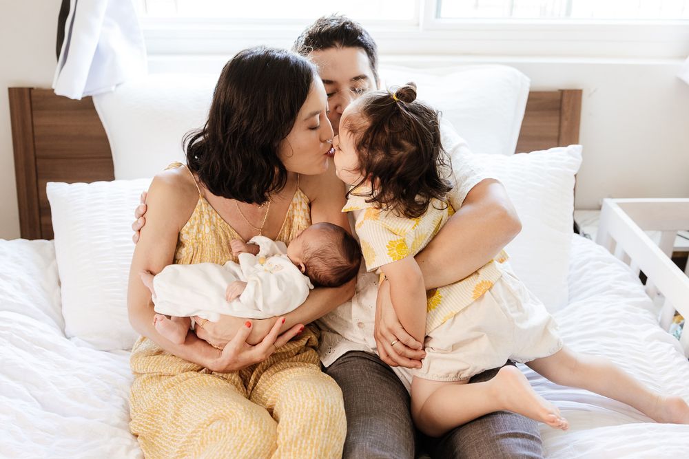 Gentle family snuggles on the bed, as mom kisses big sibling while holding baby - San Jose newborn photographer
