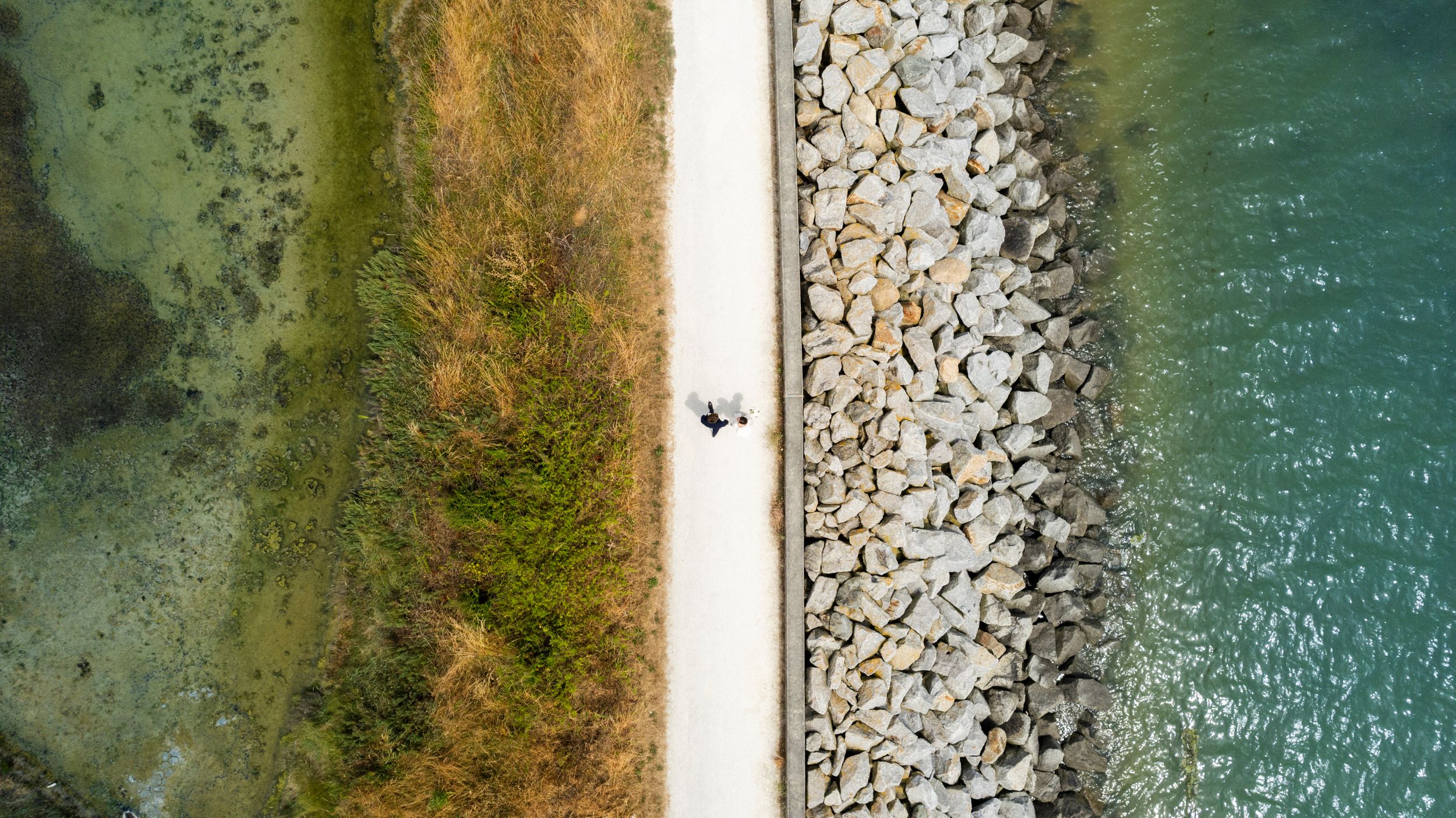 Aerial view of a coastal path with rocks and turquoise water on either side as a person walks along the seawall.