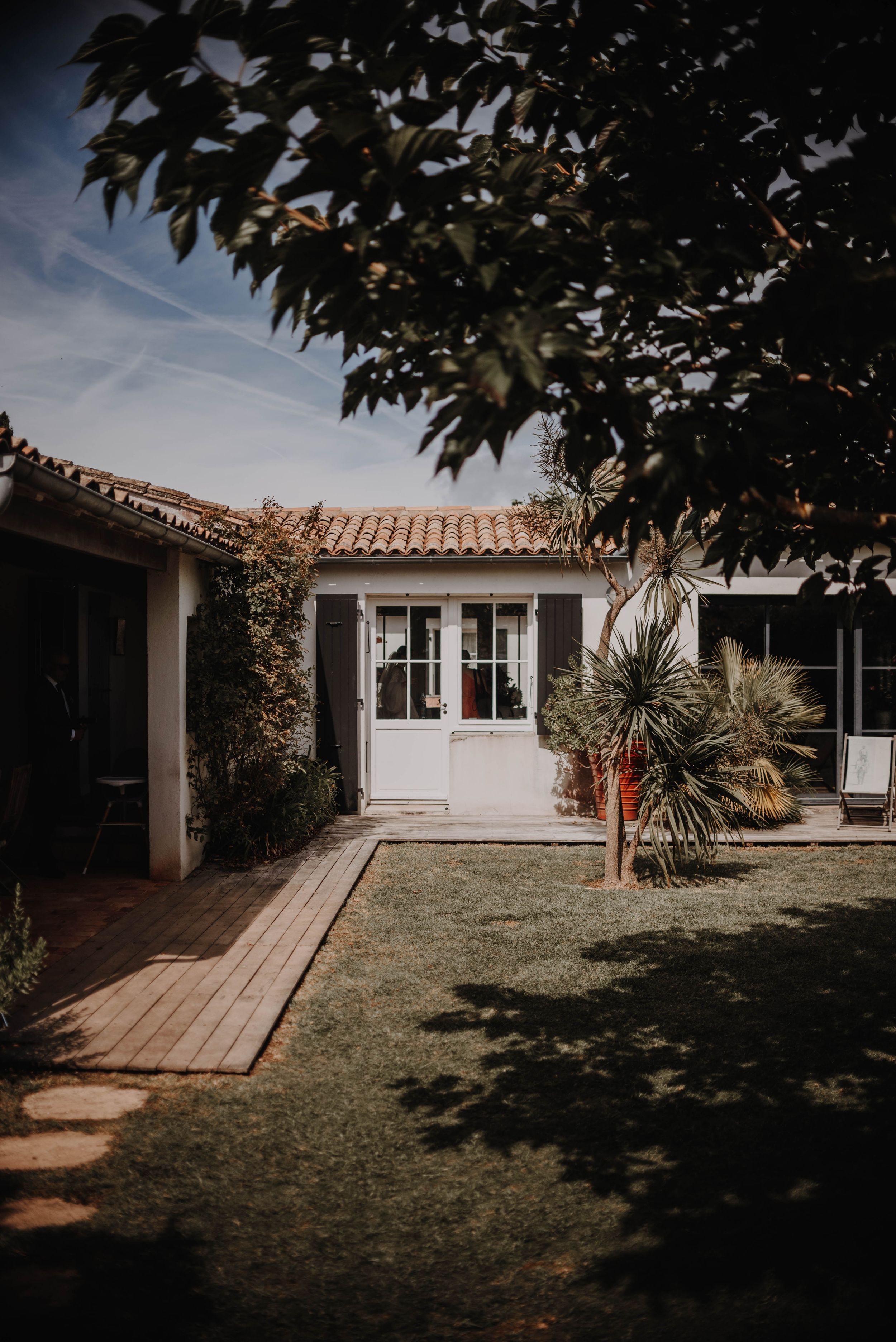 Modern white house with wooden deck and landscaped yard featuring palm trees and decorative plants in natural sunlight.