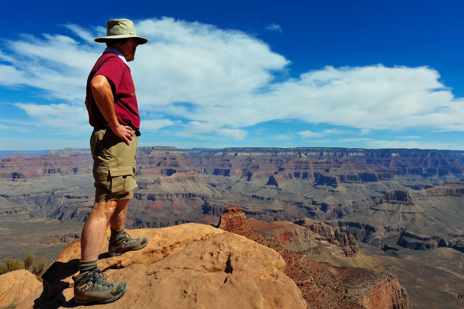 A hiker stands on a scenic overlook at the edge of the Grand Canyon with vast desert landscape stretching to the horizon.