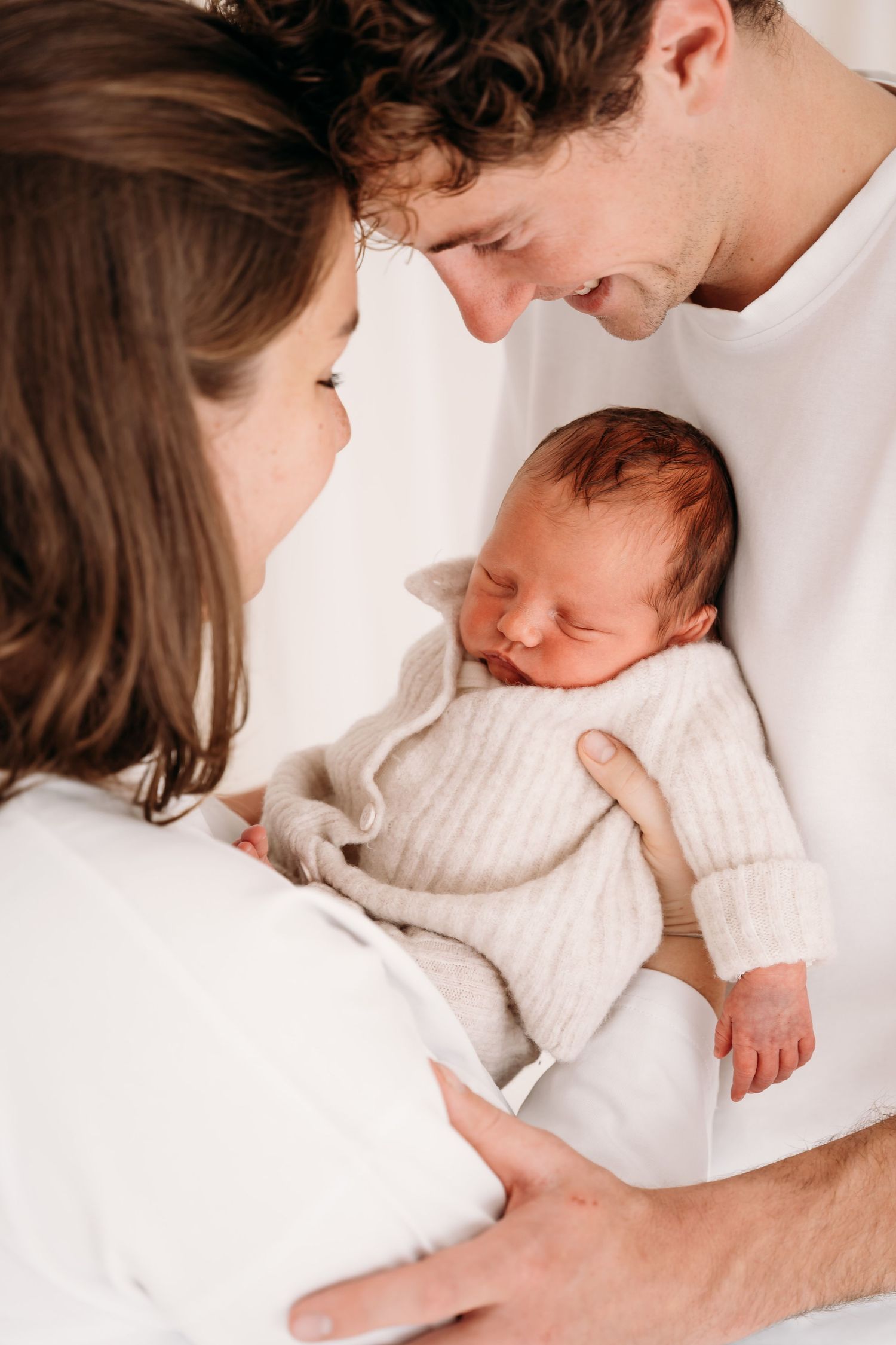 Gezin met newborn dicht bij elkaar, zachte blik en handen die de baby omsluiten.