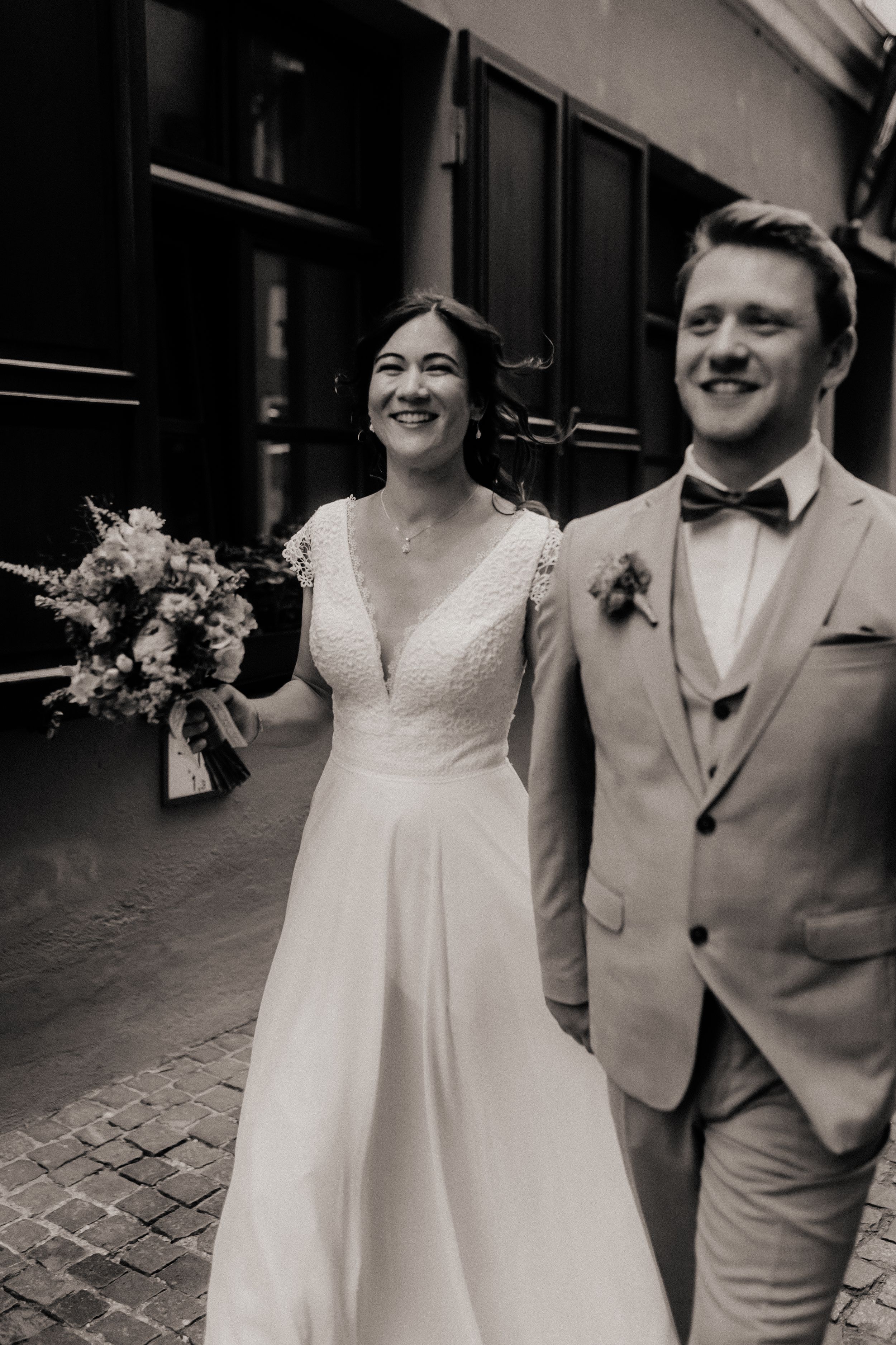 Black and white wedding photos showing a couple walking together on cobblestone street with bride holding floral bouquet.