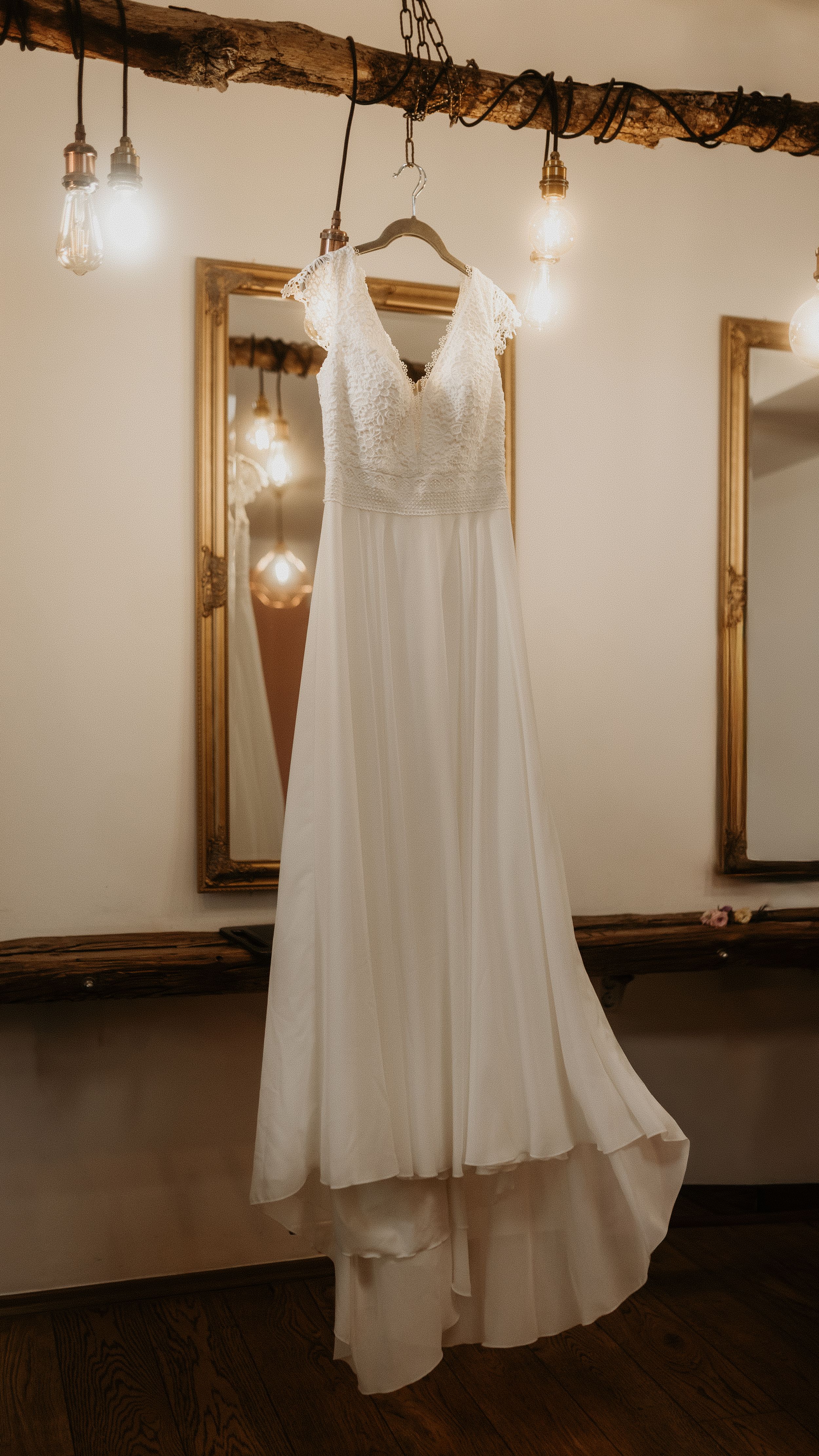 A white wedding dress hangs elegantly from a wooden hanger under pendant lights in a rustic venue setting.