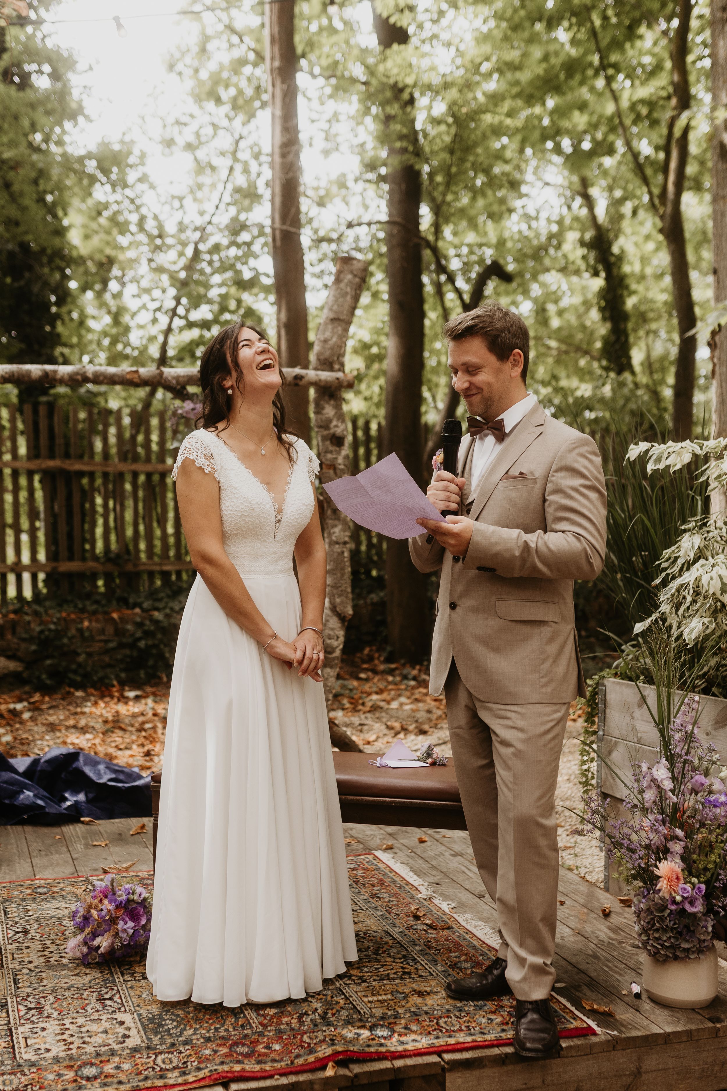 A couple shares joyful laughter during their outdoor wedding ceremony in a wooded garden setting.