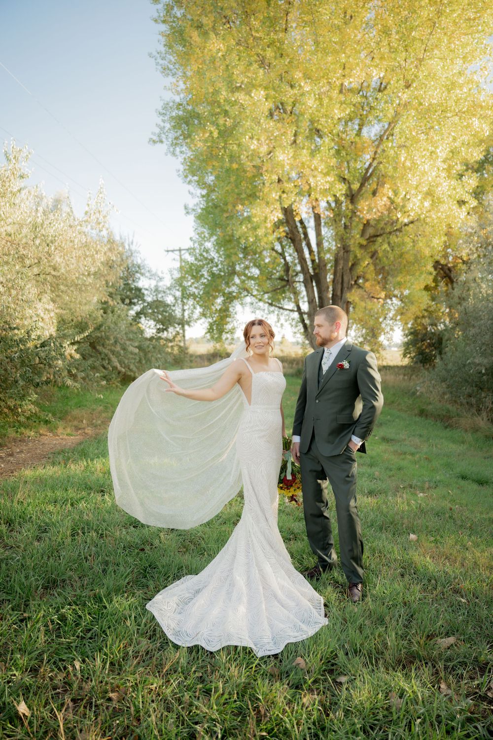 A couple poses together in a romantic autumn setting with golden trees while the bride's white dress flows in the breeze.