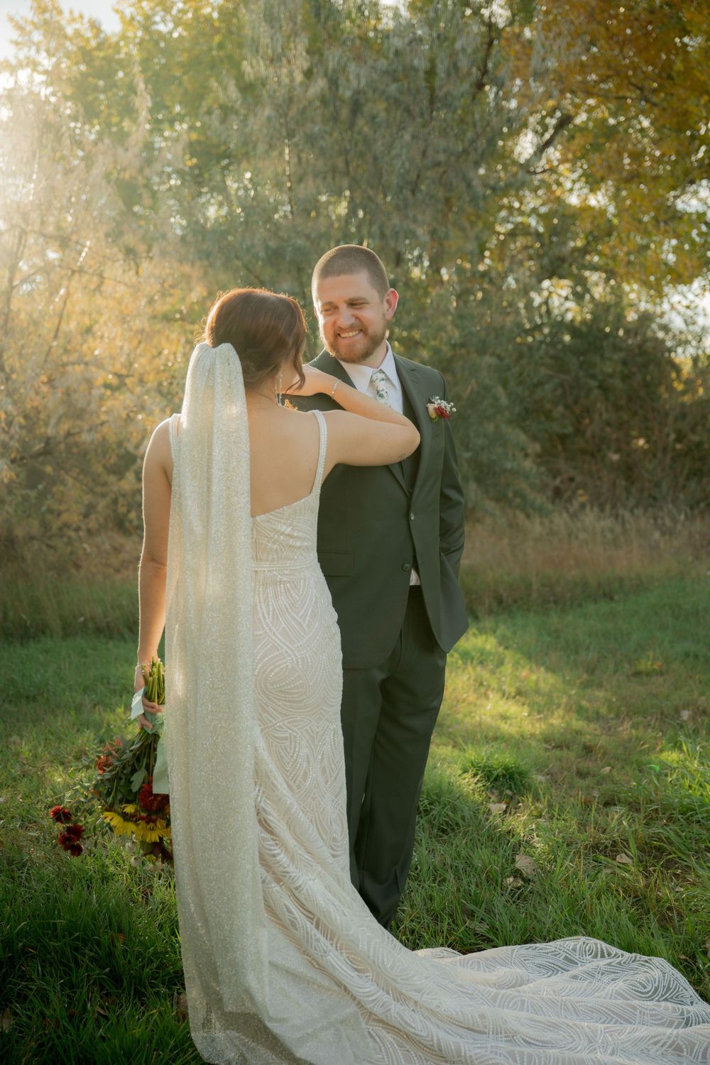A couple stands together in an autumn garden setting with golden sunlight filtering through trees behind them.