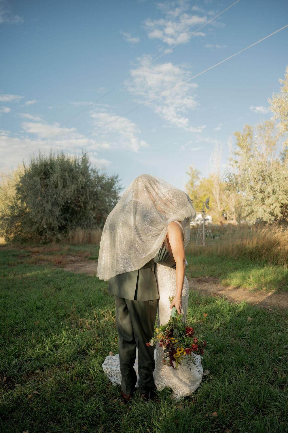 A figure in a veil and dark pants stands in a grassy field holding a bouquet of red flowers at sunset.