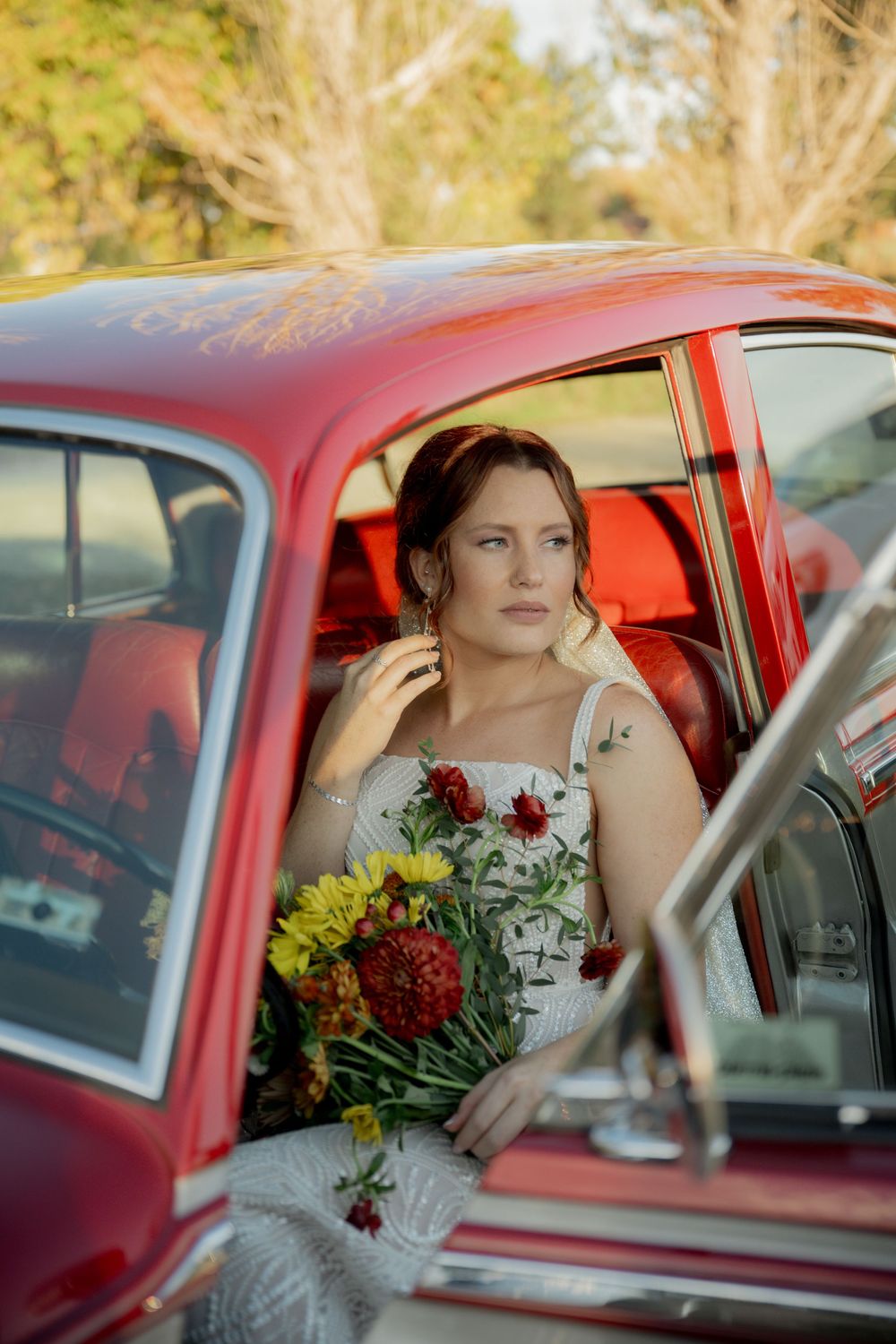 A bride in a vintage red car holds a colorful bouquet of flowers on her wedding day.