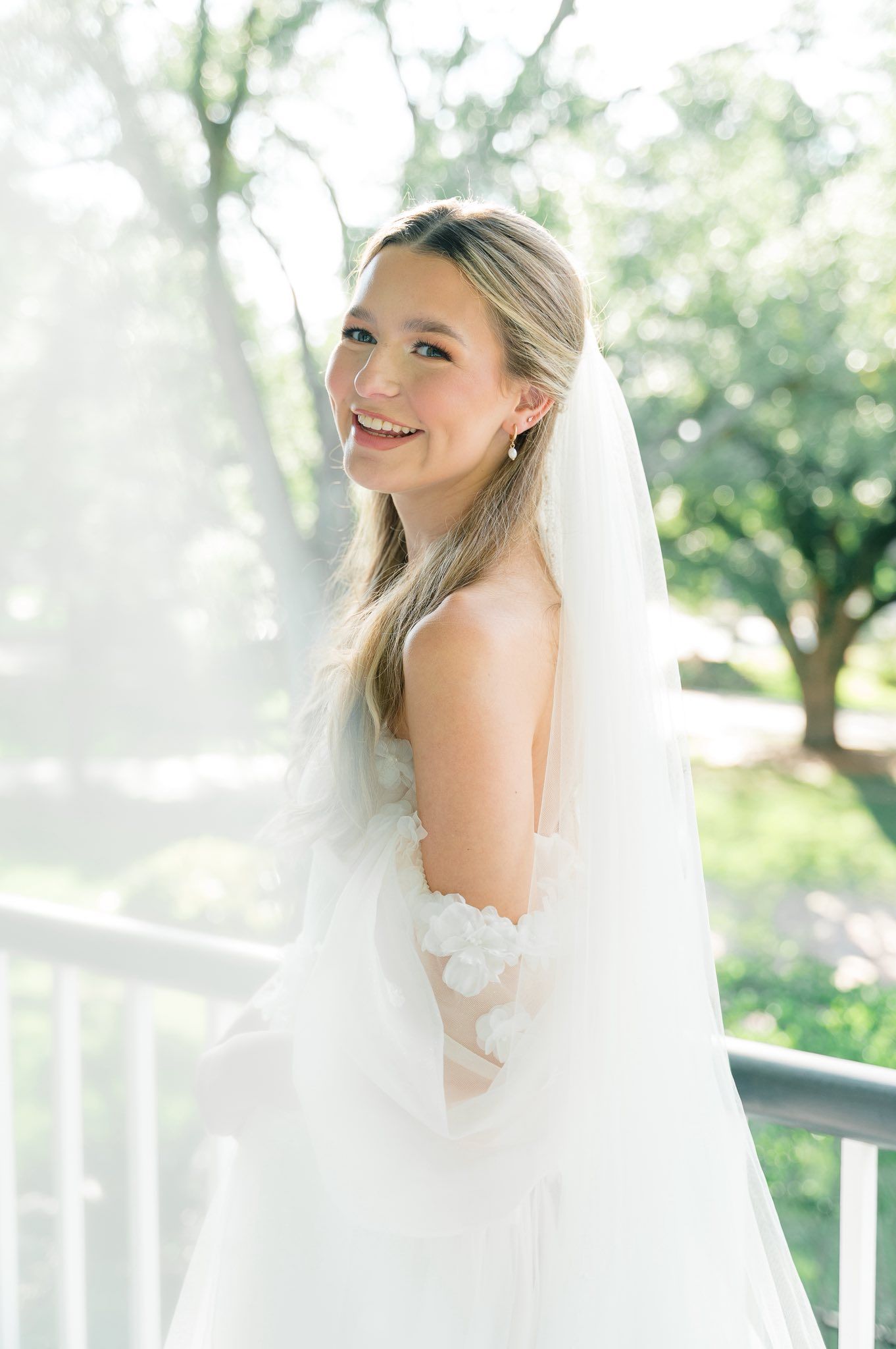 Elegant bridal portrait with flowing veil captured in soft natural light on a white porch with greenery in background.