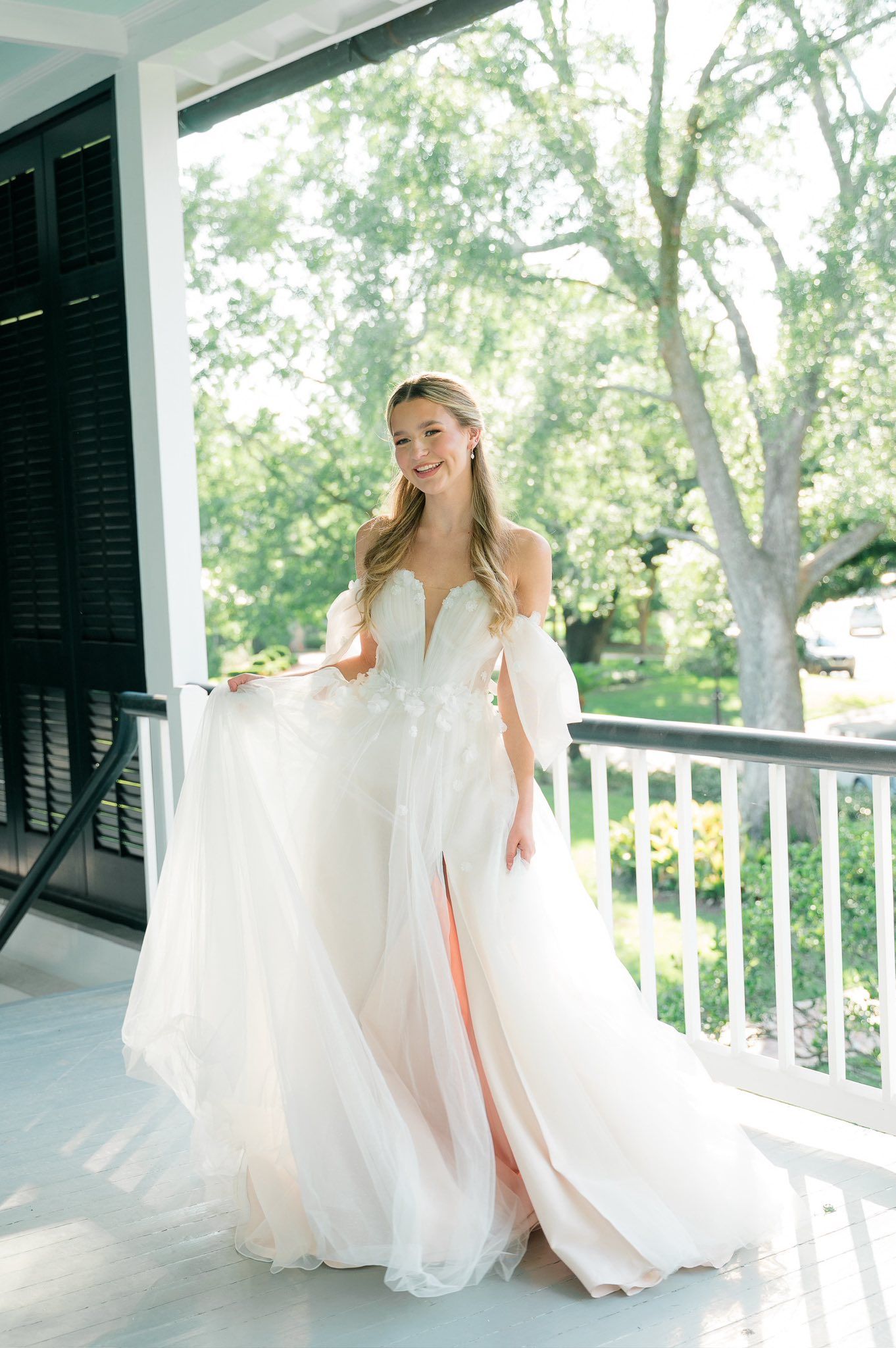 Two elegant bridesmaids in flowing pink and white dresses twirl together on a sunlit porch.