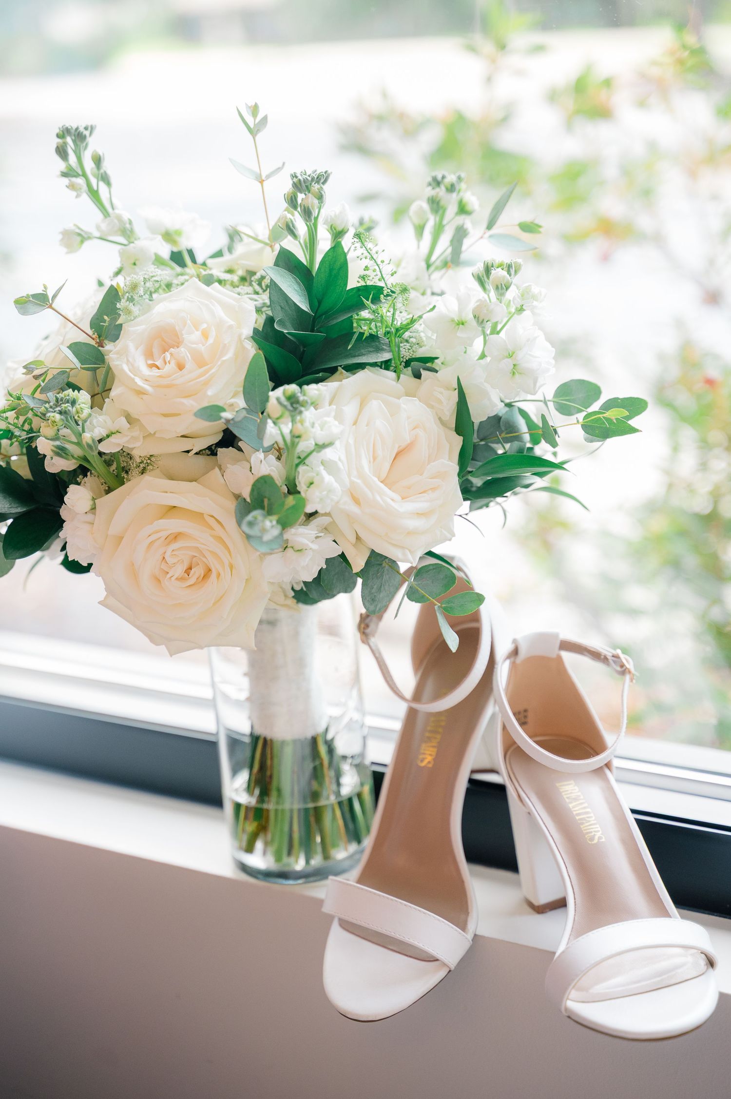 White wedding shoes and delicate cream roses in glass vases sit elegantly on a windowsill.
