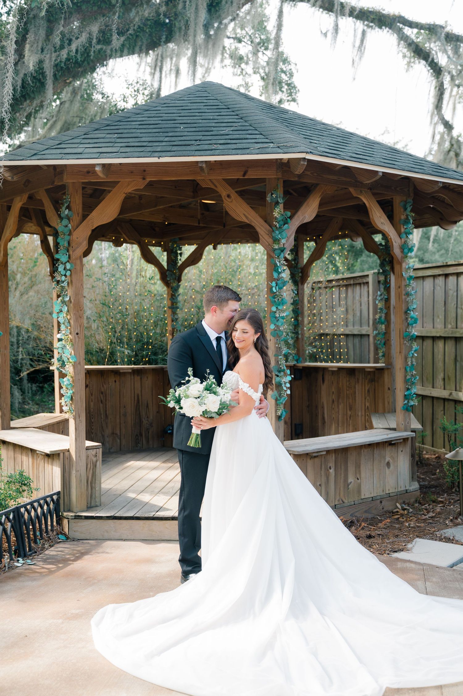 A romantic wedding couple shares a tender moment under rustic wooden gazebos surrounded by Spanish moss and greenery.