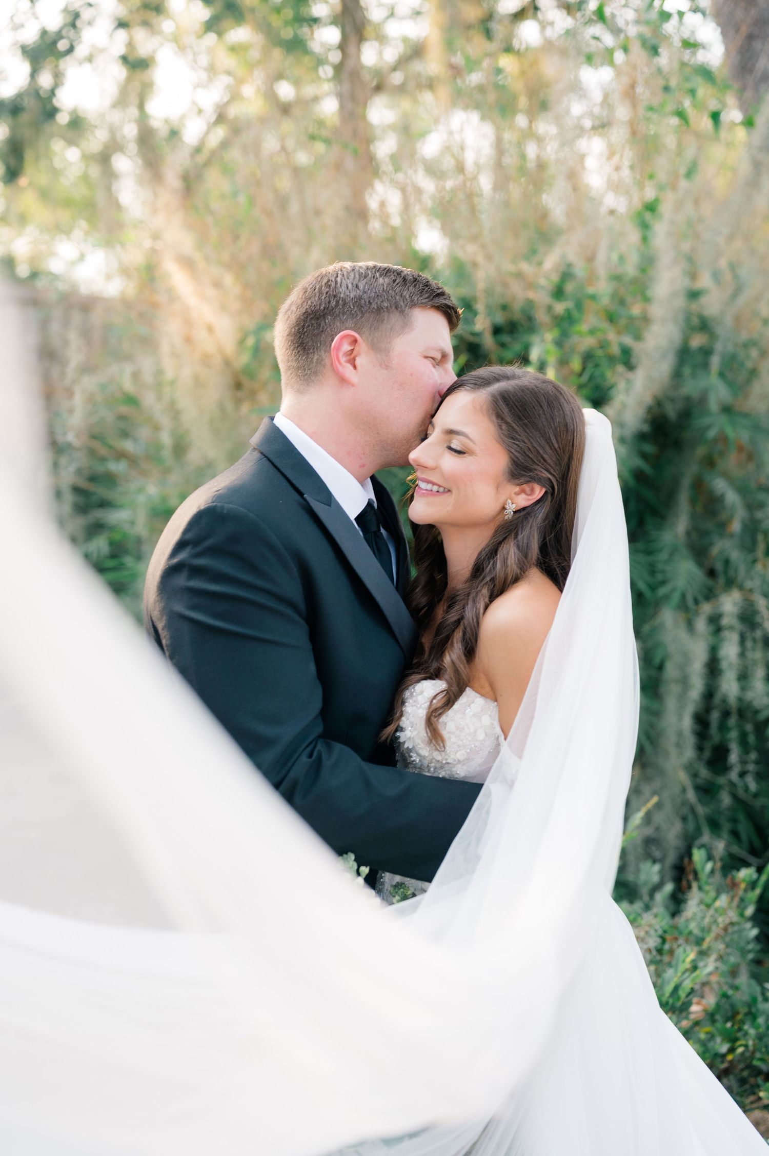 A romantic wedding portrait showing a couple embracing outdoors with a flowing white veil in natural lighting.