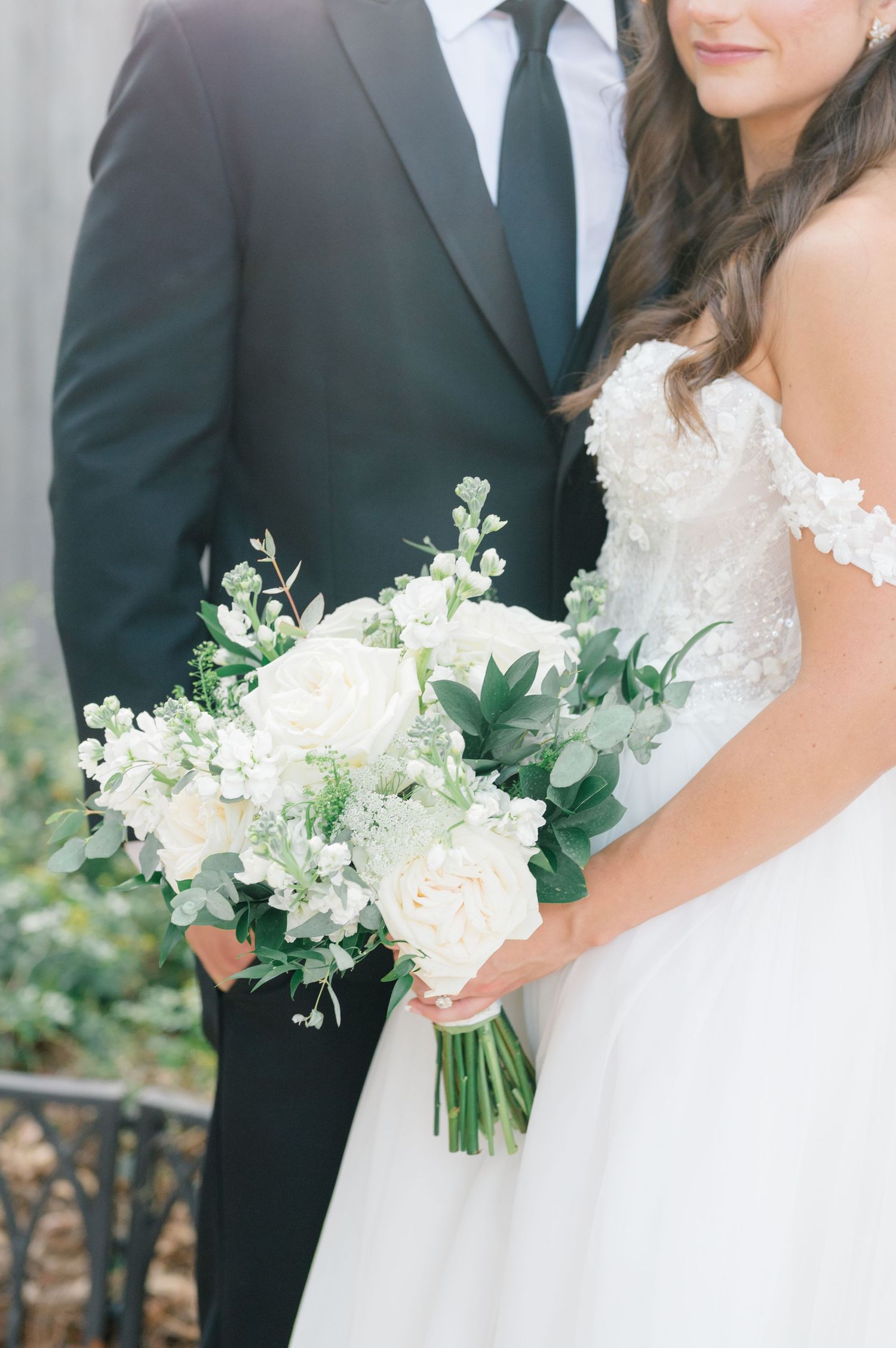 Elegant wedding couple in formal attire with white floral bouquet featuring delicate greenery and blooms.