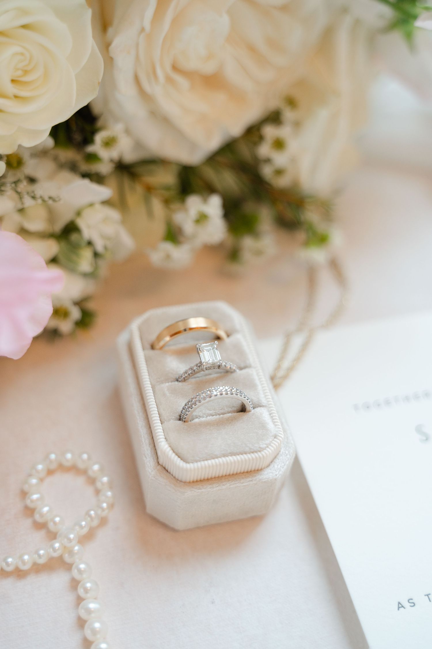 Wedding ring displayed in a white velvet box surrounded by white roses and pearl jewelry on a light background.